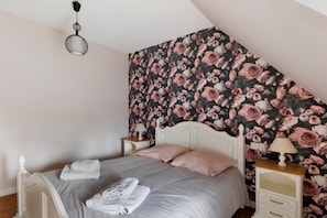 Un dormitorio con techo inclinado, una cama con ropa de cama blanca, dos mesitas de noche con lámparas y un papel tapiz floral.