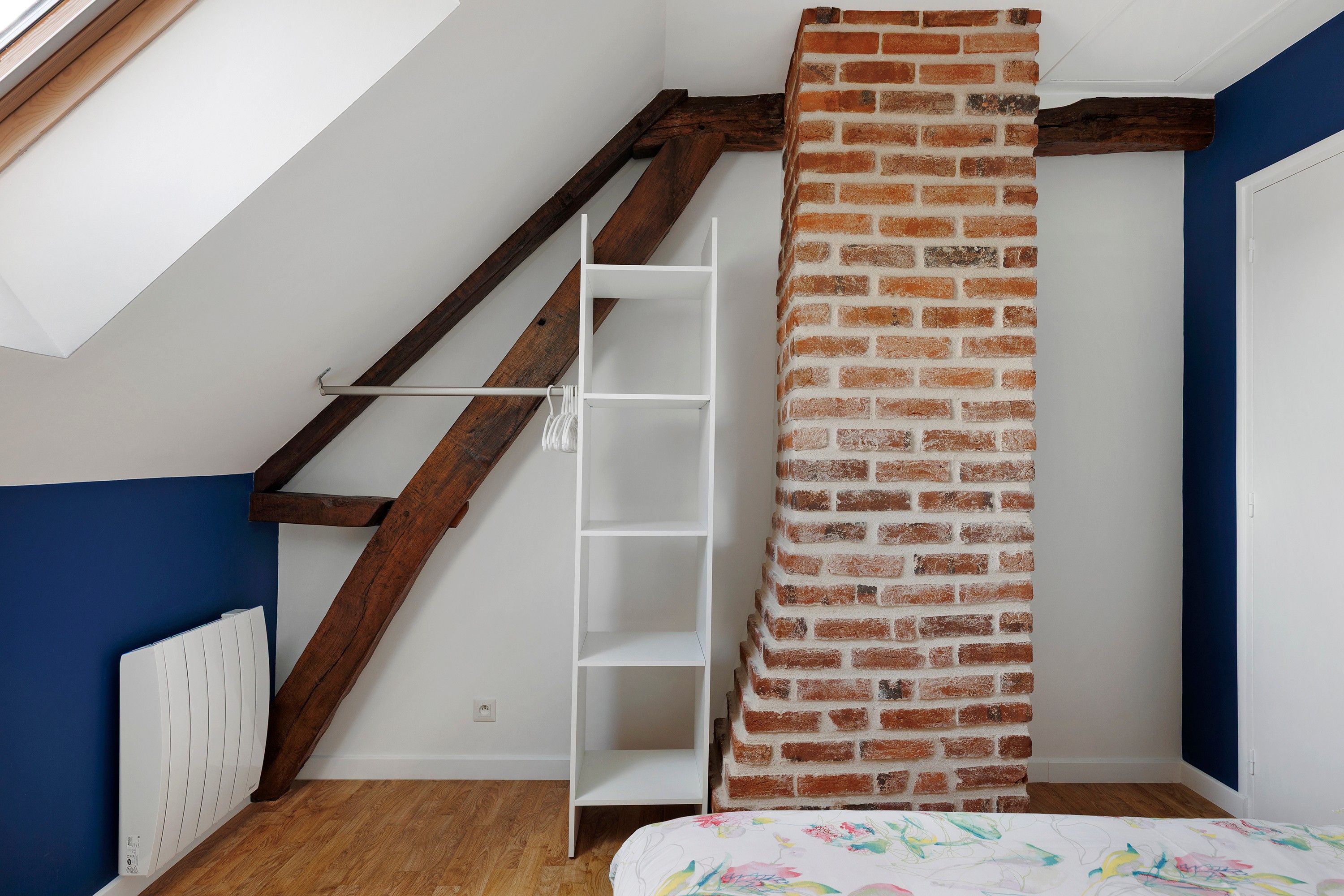 Une pièce avec un plafond en pente, un escalier en bois, une étagère blanche et un mur en briques.