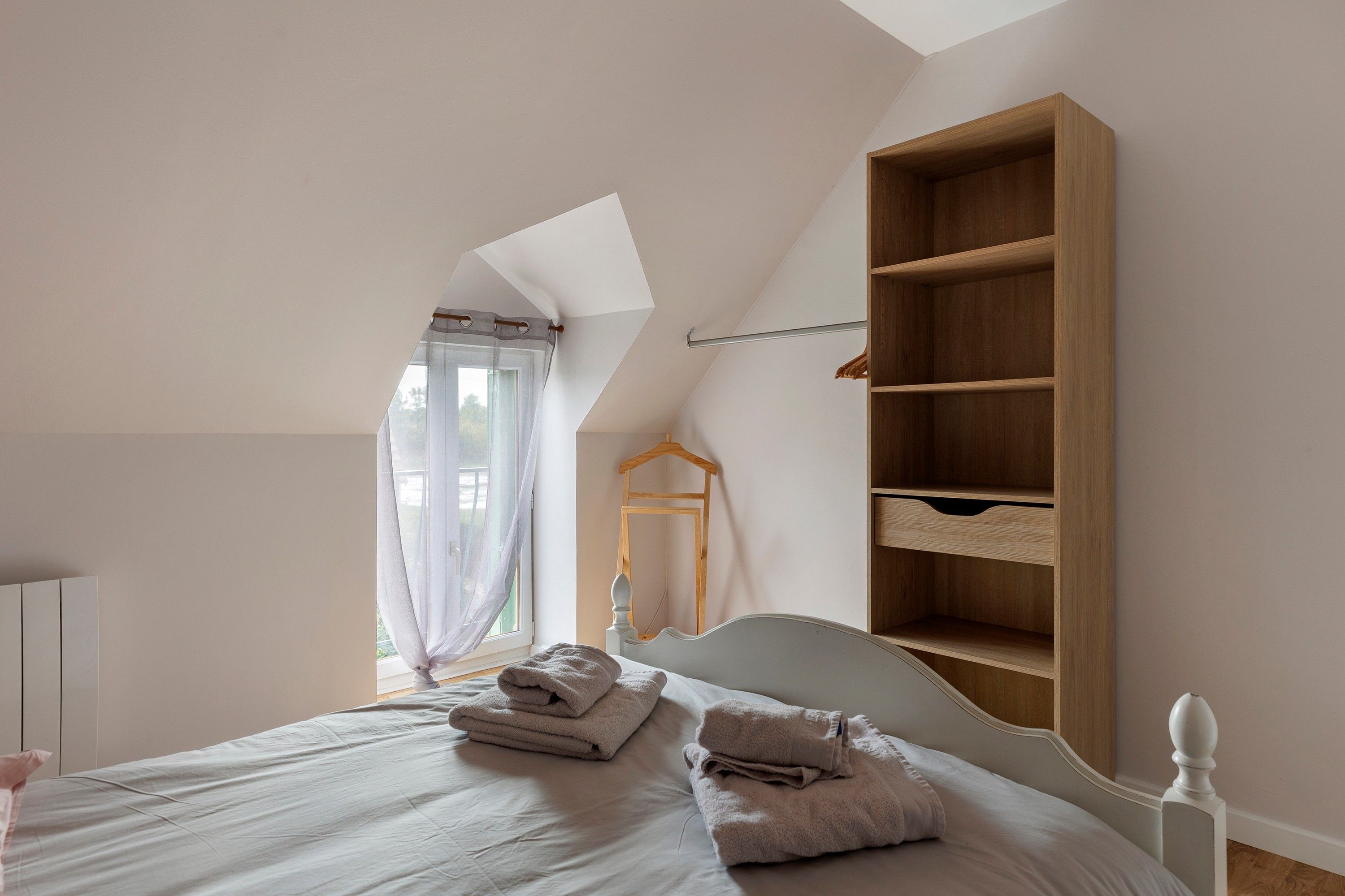 Een slaapkamer met een schuine kap, een bed met hoofdeinde, een houten garderobe en een raam met gordijnen.