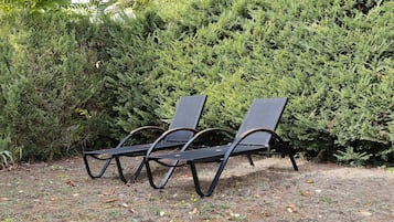 Twee zwarte lounge stoelen met metalen frame en kussens, geplaatst op een grindpad met herfstbladeren, omgeven door dichte groene struikgewas.