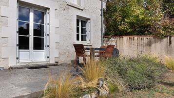 A two-story building with a stone facade, a small patio with a table and chairs, and a garden area with various plants.