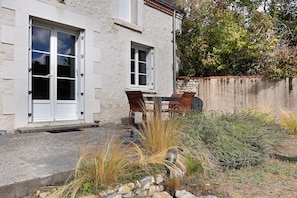 A two-story building with a stone facade, a small patio with a table and chairs, and a garden area with various plants.