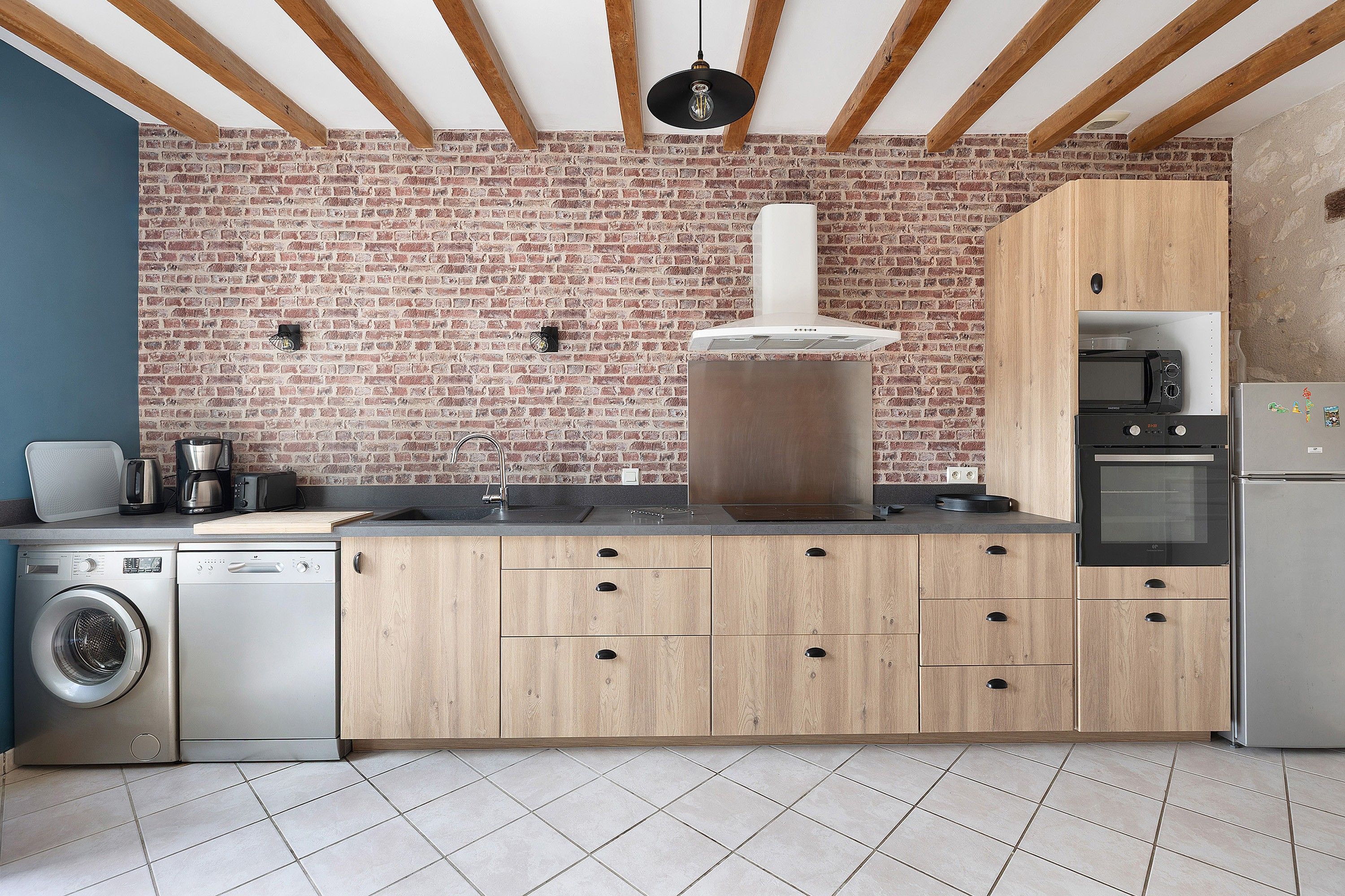 Une cuisine avec un mur en briques, des meubles en bois, un four encastré et un lave-linge.