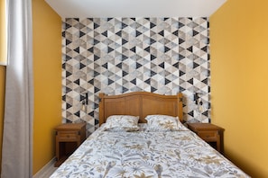 Une chambre à coucher avec un papier peint à motifs géométriques, une tête de lit en bois, une couvre-lit à motifs floraux et deux tables de chevet avec des lampes.