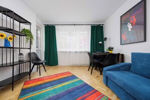 A modern living room with a blue sofa, a colorful striped rug, a dining area with a table and chairs, a bookshelf with plants, and a large window with green curtains. - Warsaw | Złota 11 | Comfortable Apartment | Downtown (Warszawa)