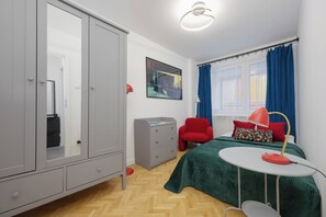 A modern bedroom with a wardrobe, a desk, a bed with a green bedspread, a red chair, and a window with blue curtains. - Warsaw | Złota 11 | Comfortable Apartment | Downtown (Warszawa)