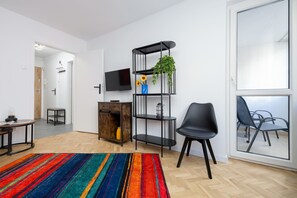 Un salon moderne avec un téléviseur, une étagère noire à plusieurs niveaux agrémentée d’une plante, un fauteuil noir et un tapis rayé aux couleurs vives.
