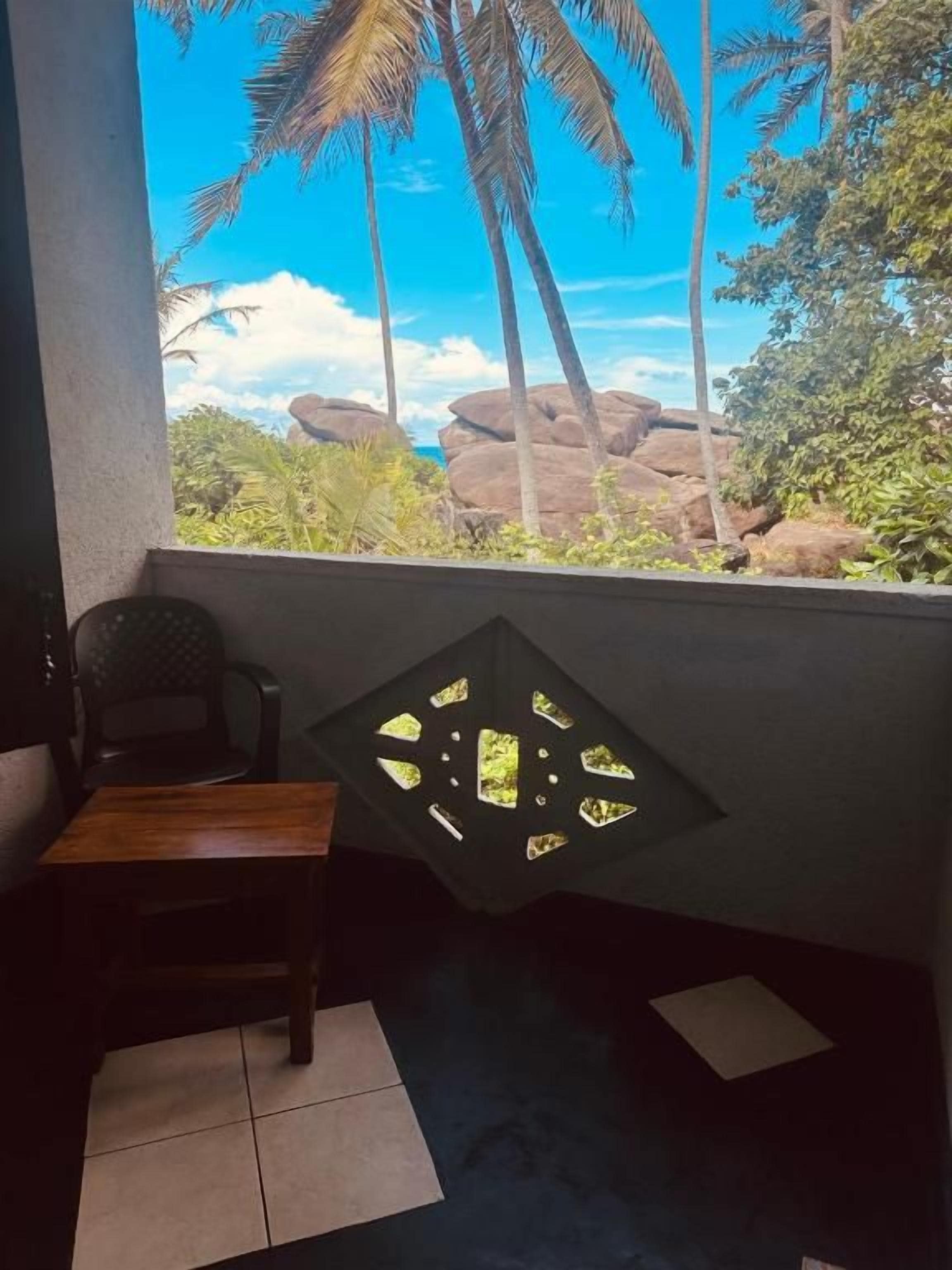 A room with a view of palm trees and rocks, a table, and a chair.