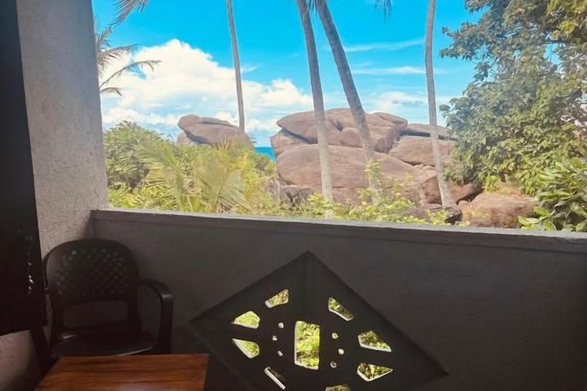 A room with a view of palm trees and rocks, a table, and a chair.