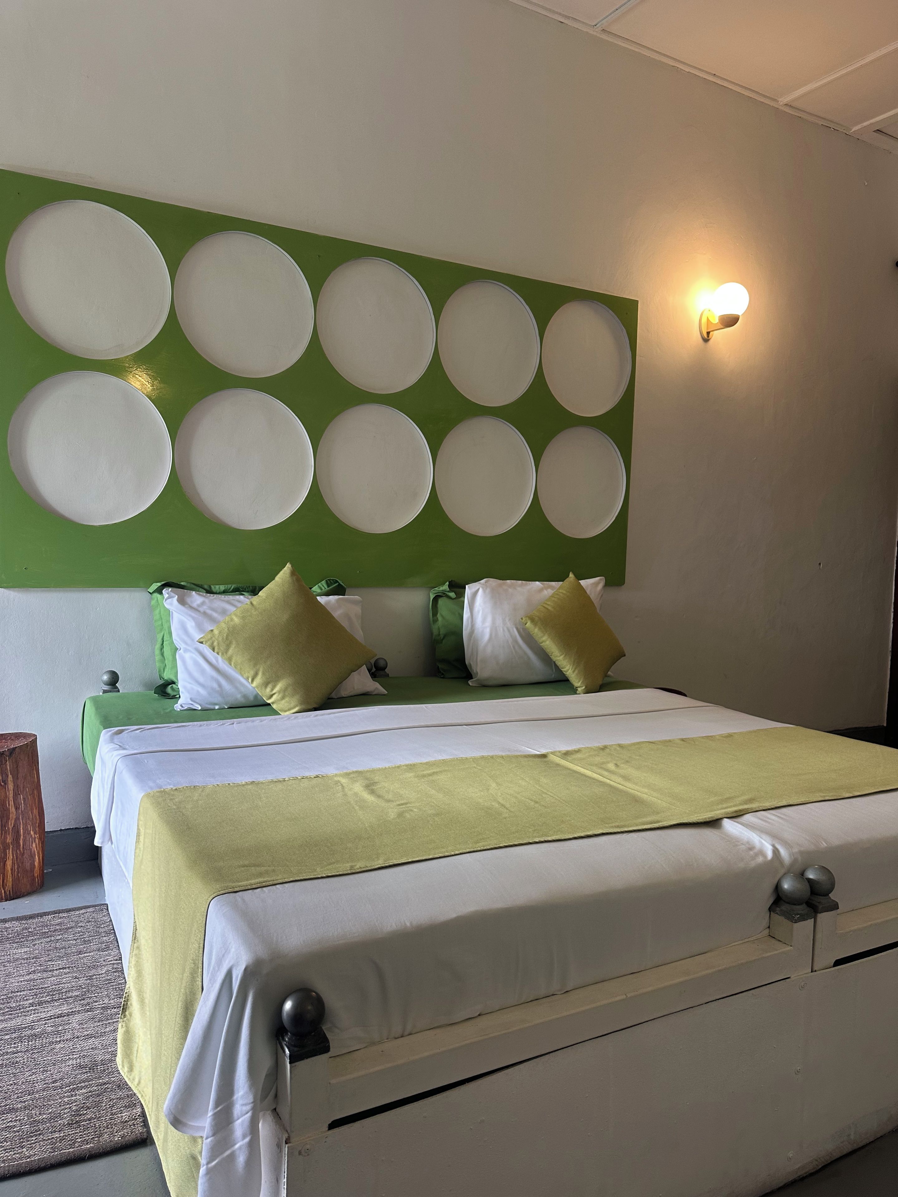 A neatly made bed with a green and white bedspread, a headboard with circular designs, and a wall-mounted light fixture.