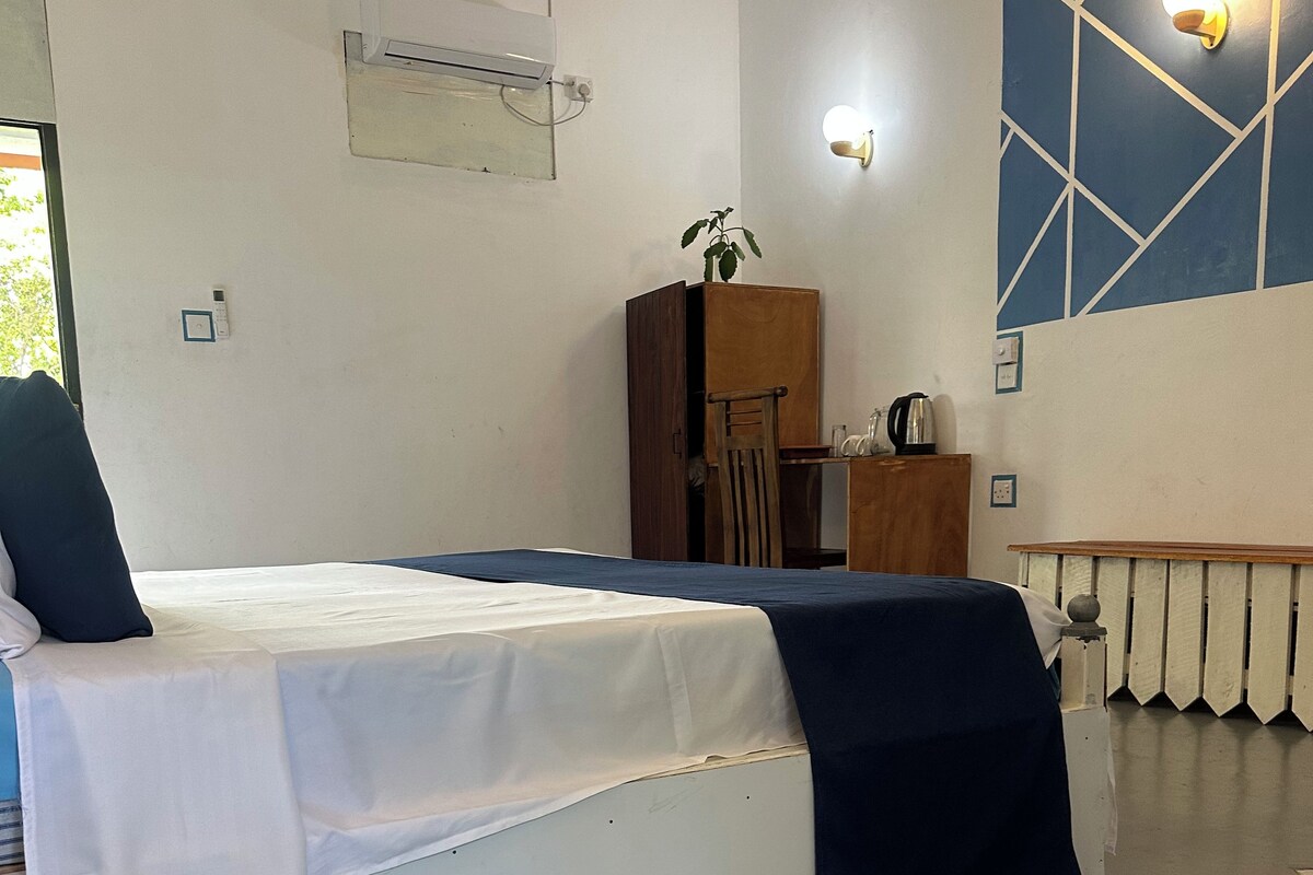 A single bed with a white and blue bedspread, a wooden wardrobe, a small table with a kettle, and a wall-mounted light fixture.