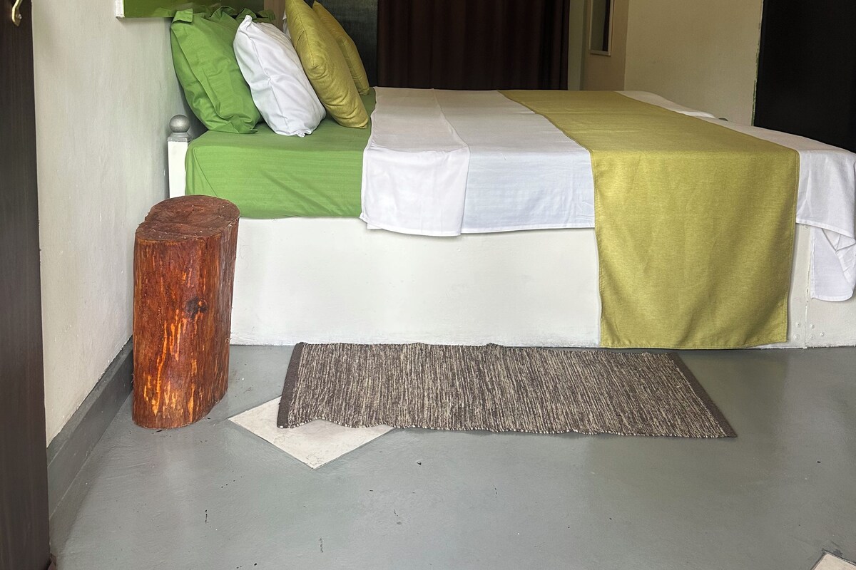 A hotel room with a bed, a green and white bedspread, a brown curtain, a wooden stool, a rug, and a wall with circular decorations.