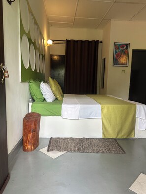 A hotel room with a bed, a green and white bedspread, a brown curtain, a wooden stool, a rug, and a wall with circular decorations. - Rock Hill Hotel  (Unawatuna)