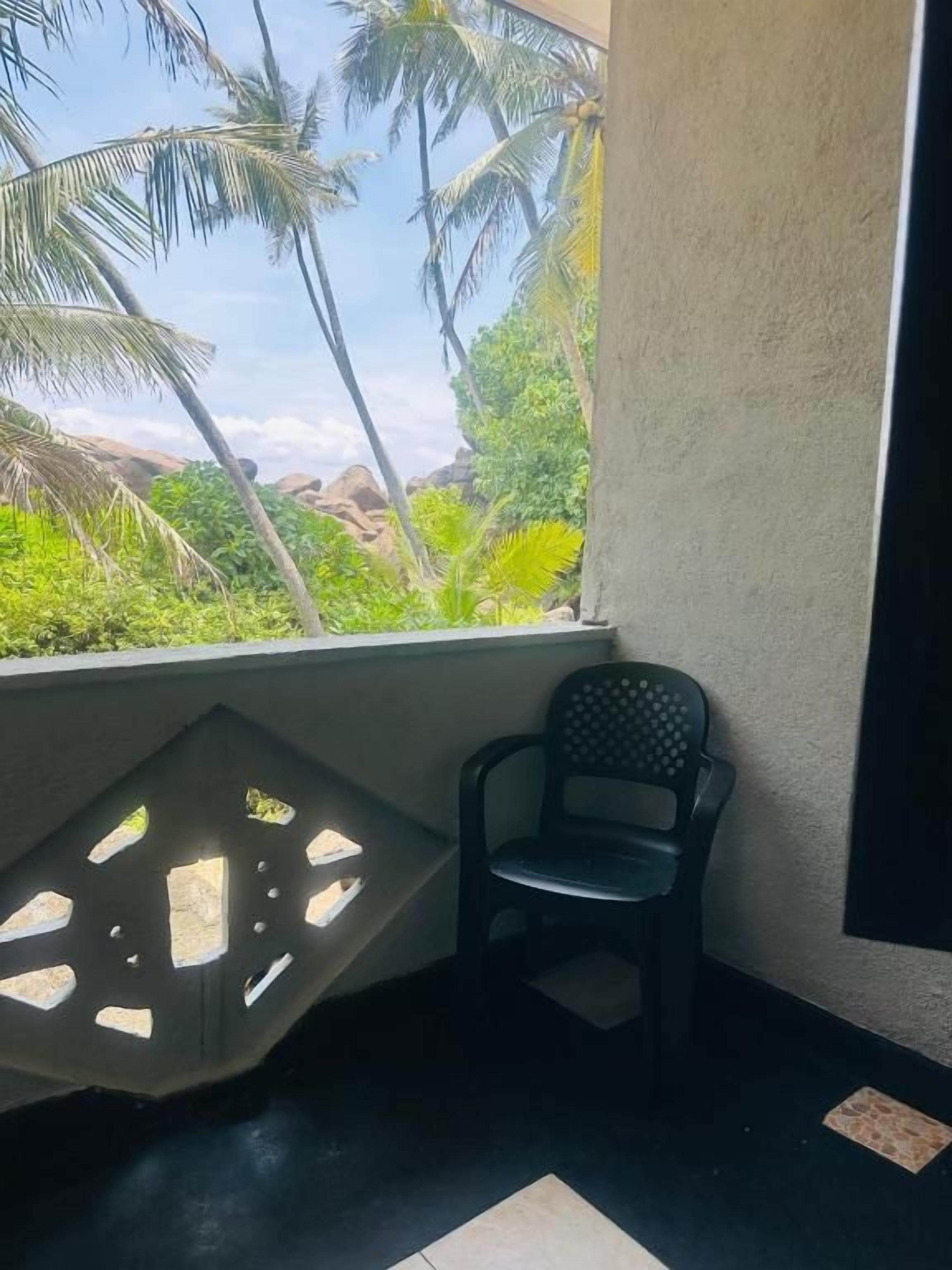Canto de um quarto com janela que dá para vegetação e palmeiras, uma cadeira de plástico e luminária fixa na parede.