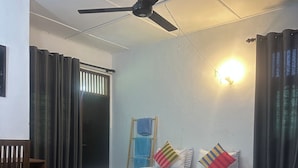Una habitación con dos camas, un ventilador de techo, una ventana con cortinas y una lámpara fijada a la pared.