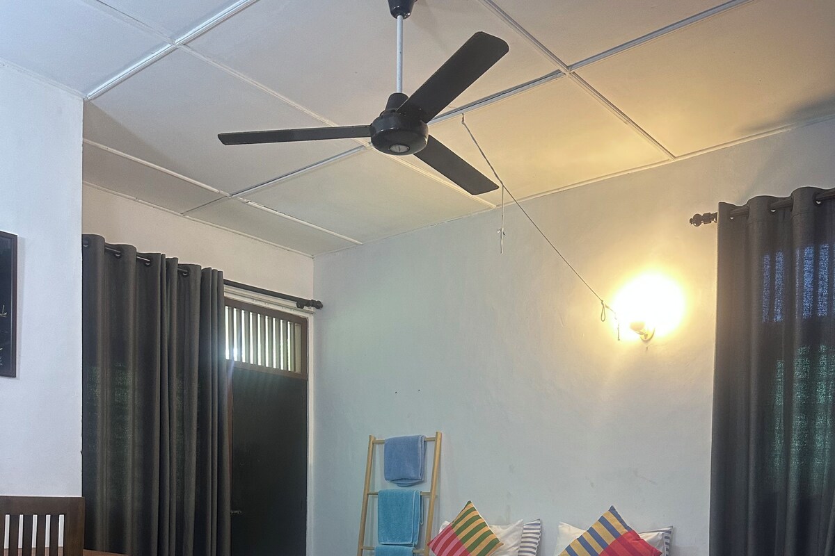 A room with two beds, a ceiling fan, a window with curtains, and a wall-mounted light.