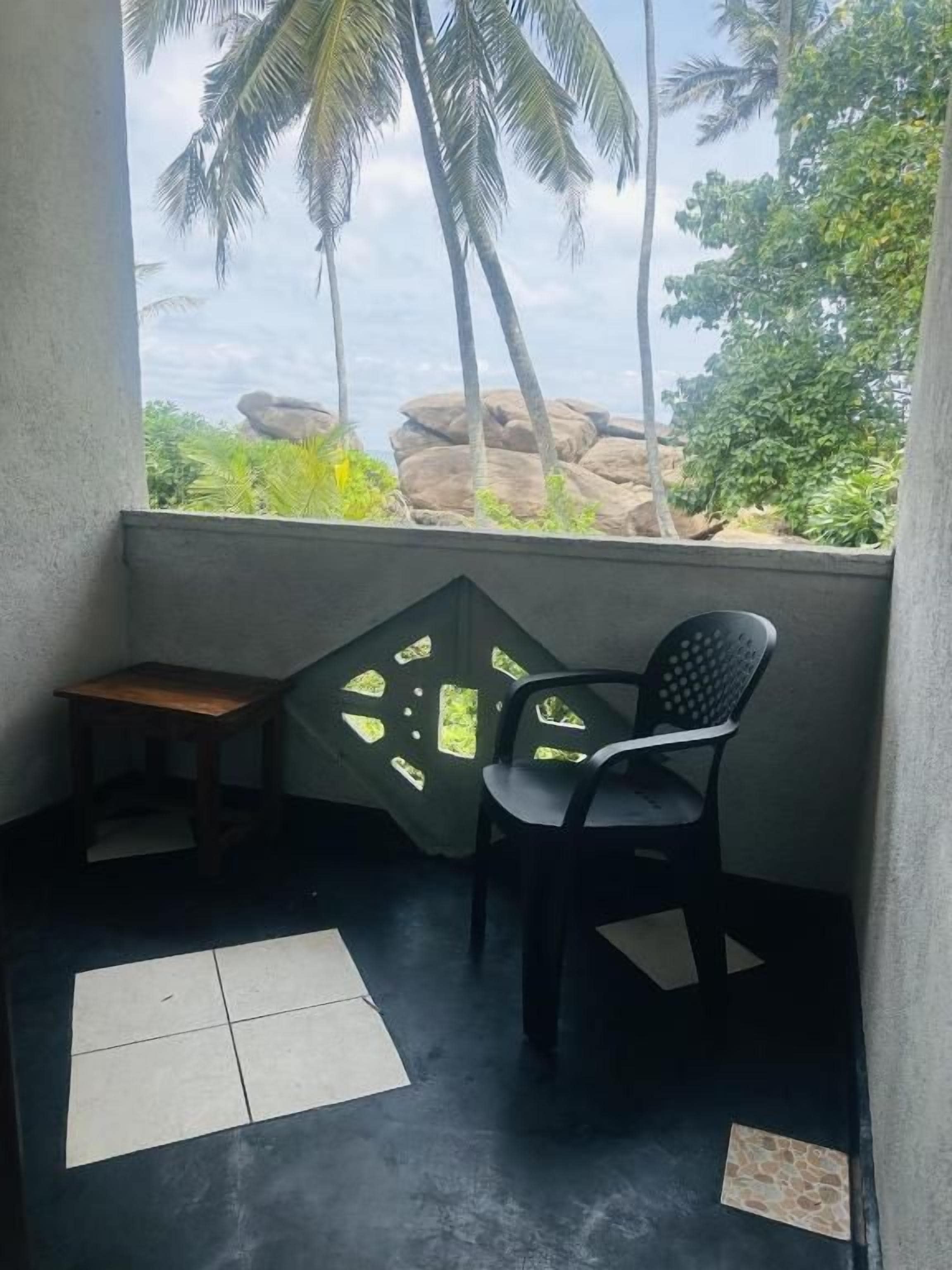 A small balcony with a chair and a table, overlooking a natural landscape with palm trees and rocks.
