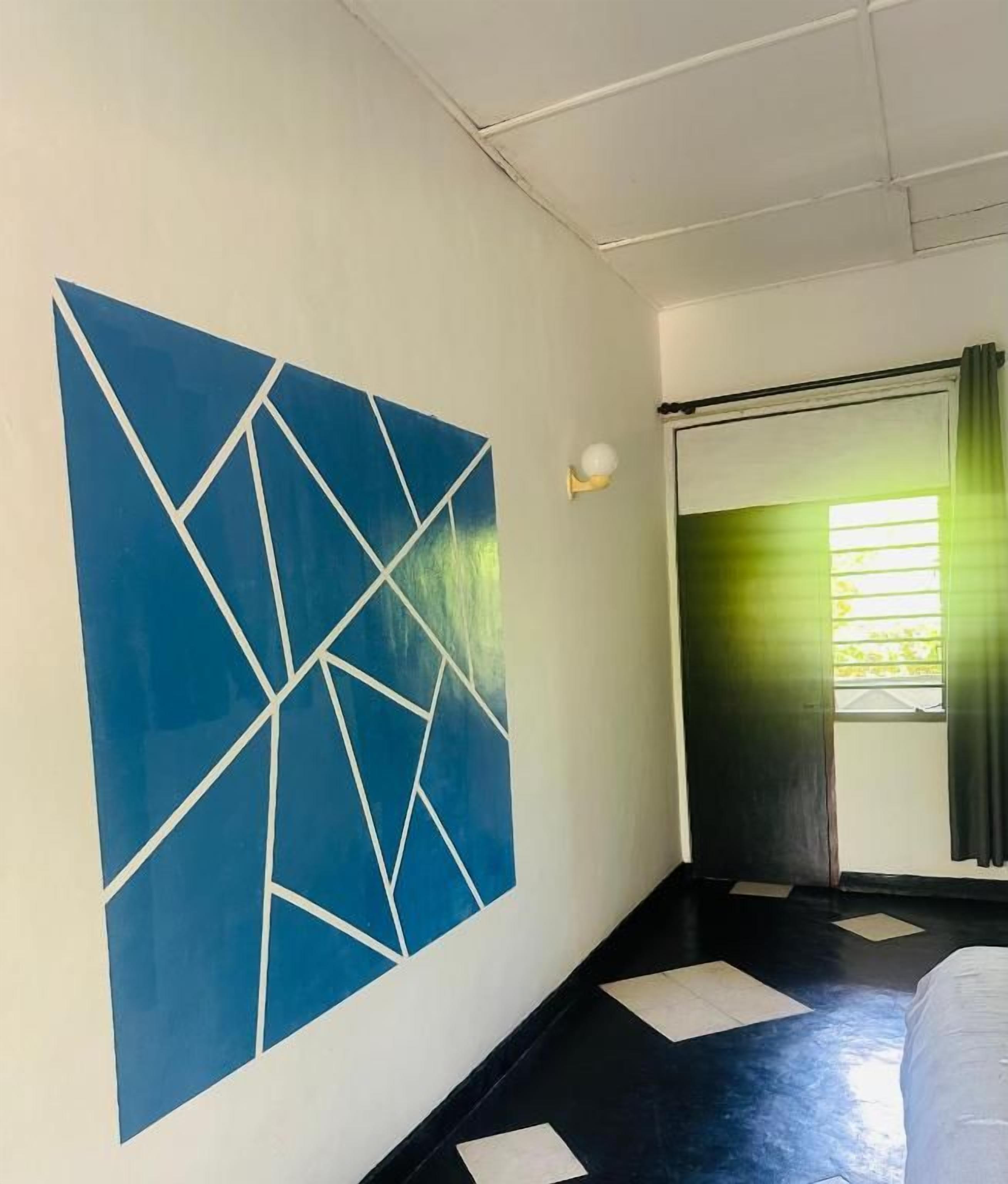A hotel room with a large geometric wall art, a bed, a window with curtains, and a door.