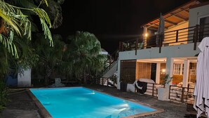 En swimmingpool med solstolar, omgiven av ett hus med en balkong och en trappa.