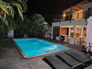 Une piscine entourée de chaises longues, elle-même bordée d’une maison dotée d’un balcon et d’un escalier.