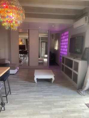 Un salon moderne avec un plancher en bois, un banc blanc, un coin repas avec des chaises et un coin cuisine équipé d’un téléviseur et de placards.