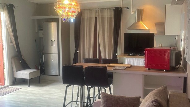 Ett vardagsrum med en soffa, en matplats med ett bord och stolar, ett kök med en spis och en röd köksapparat samt en kristallkrona.