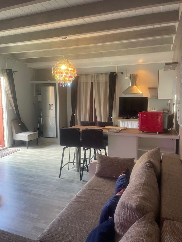 Un salon avec un canapé, un coin repas avec une table et des chaises, une cuisine avec une cuisinière et un appareil électroménager rouge, et un lustre.