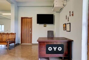 Un mostrador de recepción con un cartel que dice "OYO" en el vestíbulo de un hotel, con un banco de madera, una puerta y un televisor montado.