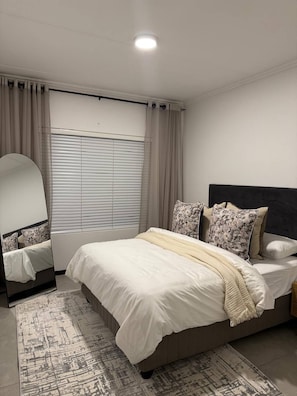 Un dormitorio con cama, espejo de cabecera, ventana con persianas y alfombra.