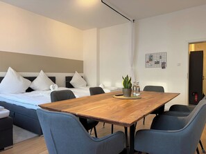 Ein modernes Hotelzimmer mit einem Holztisch, grau gepolsterten Stühlen, einem Bett mit weißer Bettwäsche und einer kleinen Topfpflanze auf dem Tisch.