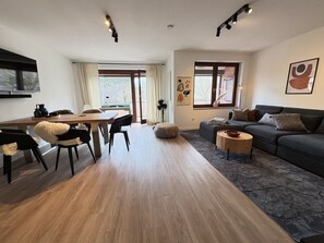 Ein modernes Wohnzimmer mit Holzboden, einem dunkelgrauen Sofa, einem runden Couchtisch und einem Essbereich mit Holztisch und Stühlen.