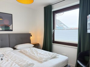 Habitación de hotel con cama, mesita de noche, lámpara, ventana con cortinas y vista a edificios.