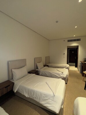 A hotel room with three beds arranged in a row, a small table, and a door leading to a hallway. - Shaza Asil Hotel (Makkah)