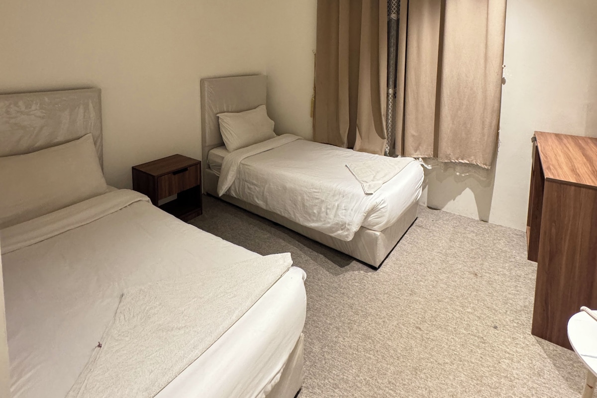 A hotel room with two beds, a wooden nightstand, a desk, and a window with curtains.