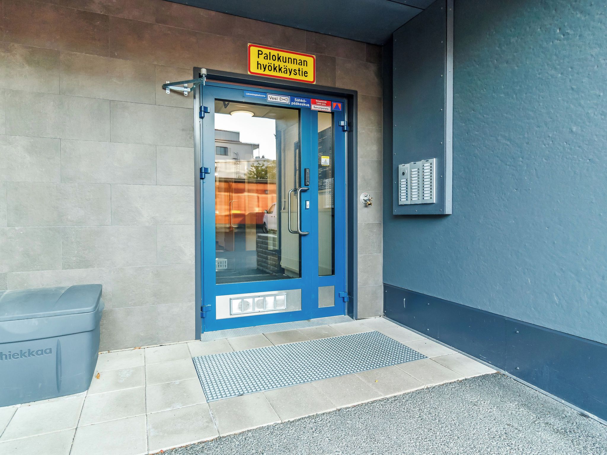 A blue door with a sign above it, a metal box on the wall, and a trash bin to the left.