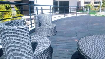 Mobiliario de mimbre para exteriores en una terraza de madera con barandilla, con vistas a un edificio con varios balcones.