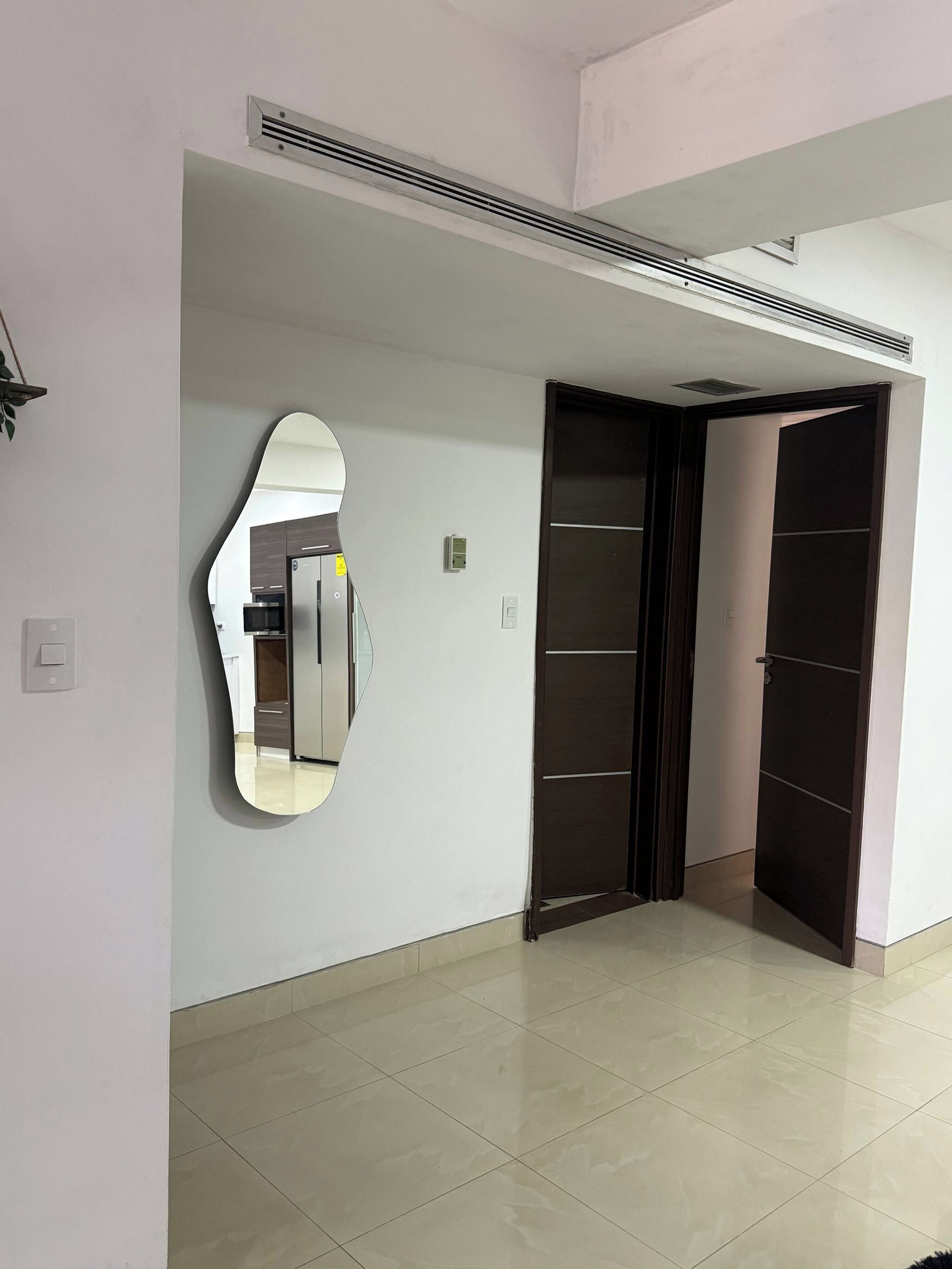 Un pasillo con un espejo grande, una puerta y una rejilla de ventilación en el techo.