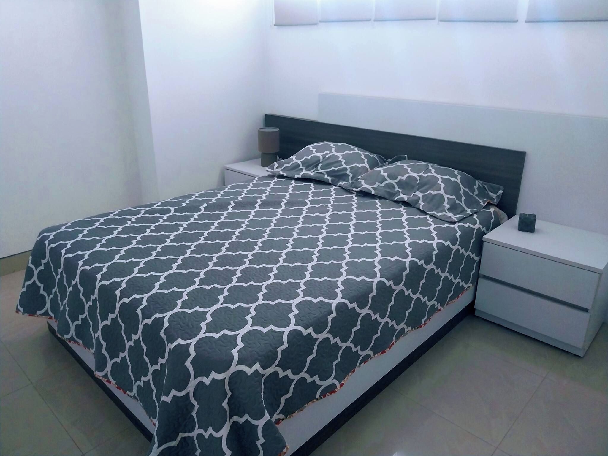 Un lit bien fait, avec une couvre-lit à motifs, un oreiller et une table de chevet sur laquelle se trouve une lampe.