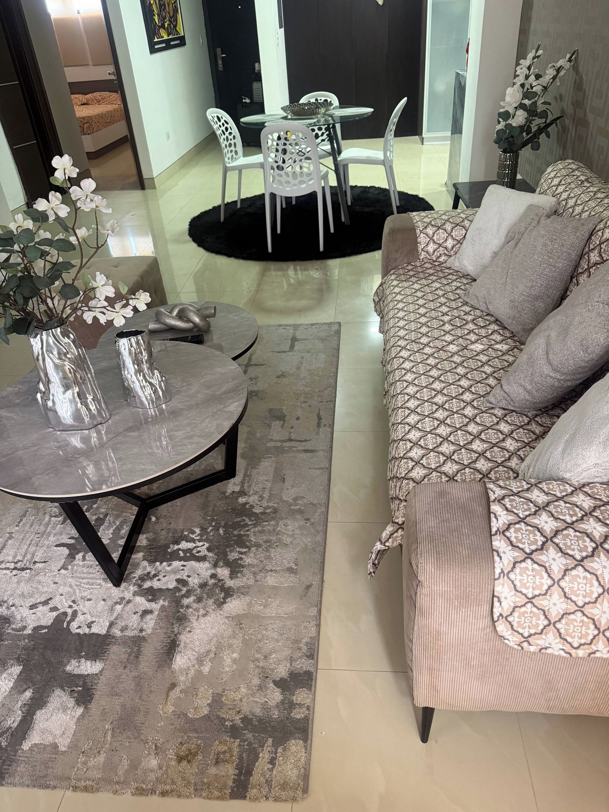 Een woonkamer met een bank, een ronde tafel en twee stoelen. Er ligt een vloerkleed en op een tafel staat een vaas met bloemen.