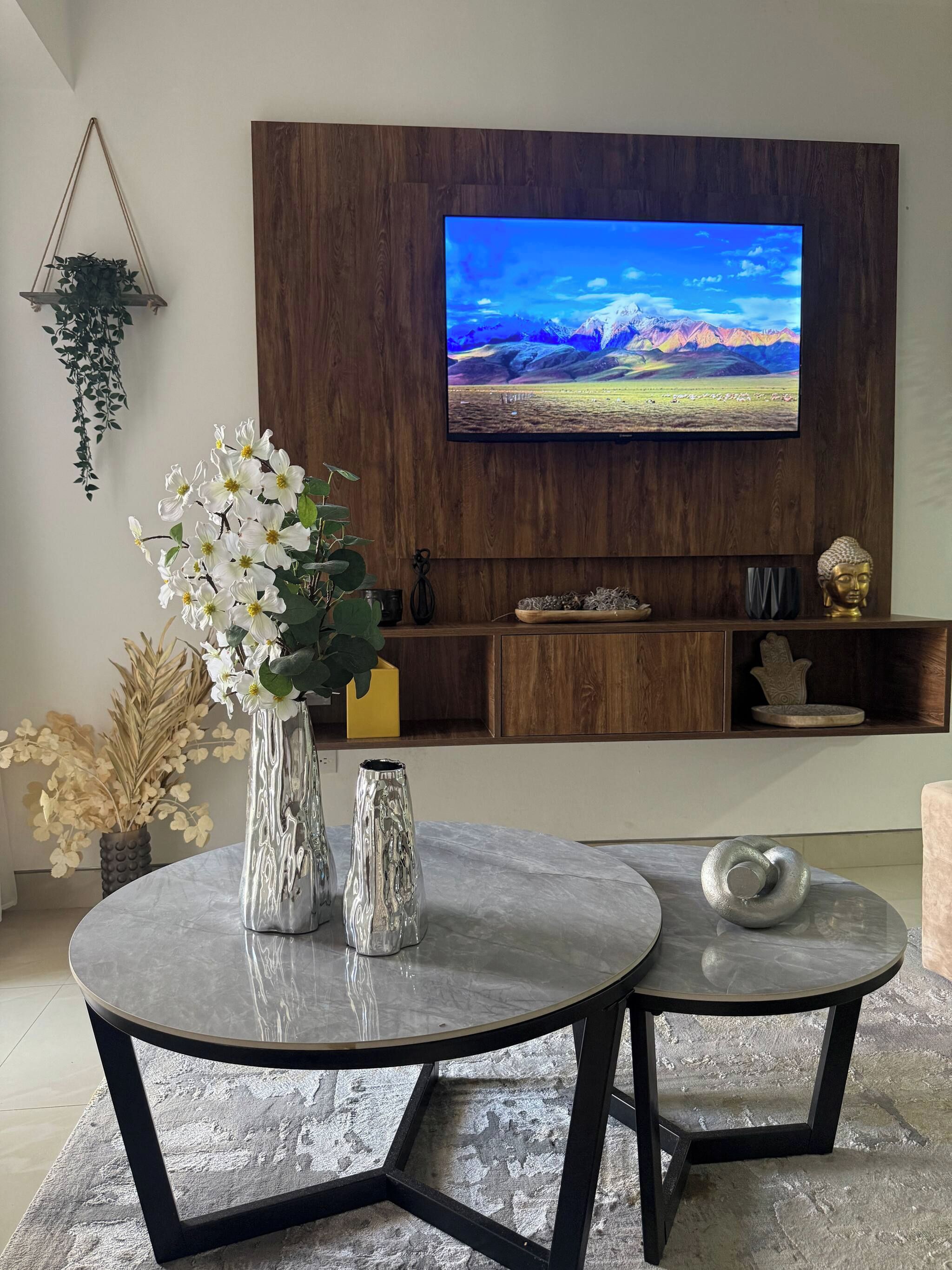 Una sala de estar moderna con un televisor de pantalla plana montado en una pared de madera, dos mesas redondas con estructura metálica y elementos decorativos como jarrones y una estatua de Buda.