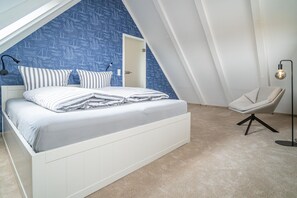 Ein Schlafzimmer mit schrägem Dach, einem Bett mit gestreifter Bettwäsche, einem Sessel und einer Stehlampe.