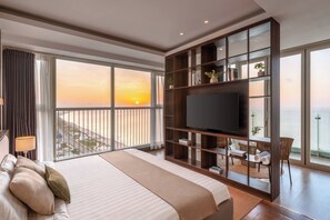 Una habitación de hotel con una cama grande, un televisor, un balcón con vistas al atardecer y una zona de comedor con muebles de mimbre.
