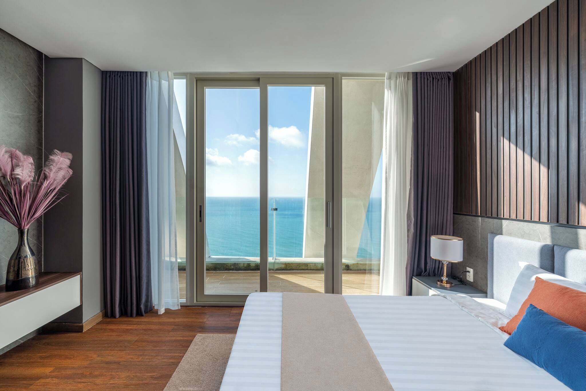 A hotel room with a bed, a nightstand, a lamp, a vase with dried flowers, and a view of the ocean through large sliding doors.