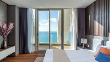 A hotel room with a bed, a nightstand, a lamp, a vase with dried flowers, and a view of the ocean through large sliding doors.