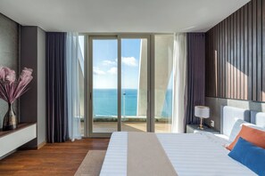 Un cuarto de hotel con una cama, una mesita de noche, una lámpara, un florero con flores secas y vista al océano a través de puertas correderas grandes.