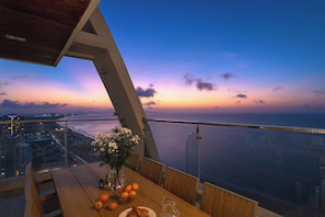 A dining table with a vase of flowers and oranges, overlooking a cityscape and a body of water during sunset. - Coho penthouse Vung Tau Luxury 3BR Oceanfront, Private Pool & Sauna (Ho Chi Minh City)