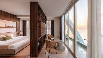Une chambre d’hôtel moderne dotée d’un grand lit, d’un coin salon avec des fauteuils, d’une petite table et offrant une vue sur la mer grâce à de grandes fenêtres.