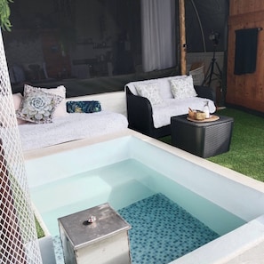 Un área de spa con una bañera de hidromasaje, dos sofás con cojines estampados, una mesita con una botella y un cuenco, y un suelo de baldosas.