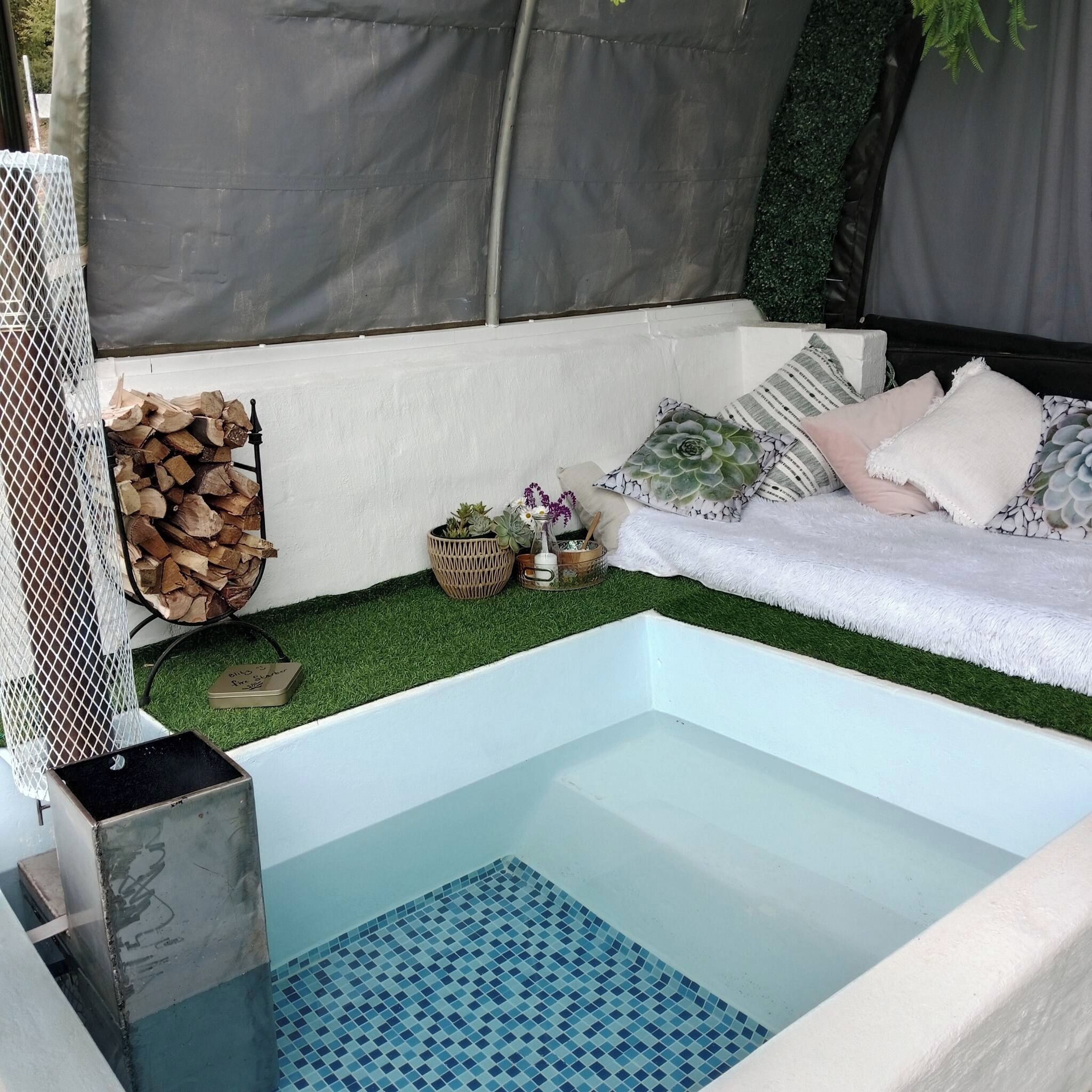 Ein kleiner Innenpool mit blau mosaikbesetztem Boden, umgeben von einer weißen Bank mit Kissen, einem Holzstapel und einer kleinen Pflanzengruppe.