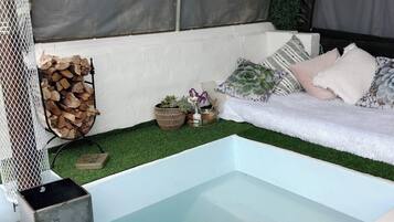 Ein kleiner Innenpool mit blau mosaikbesetztem Boden, umgeben von einer weißen Bank mit Kissen, einem Holzstapel und einer kleinen Pflanzengruppe.